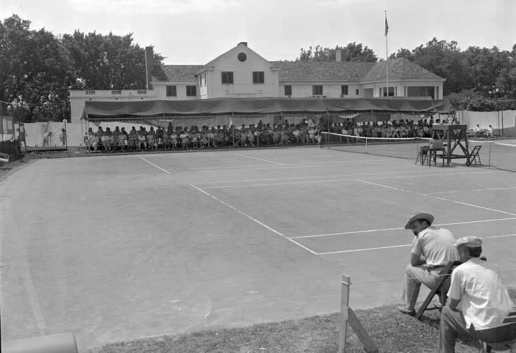 Two tennis courts are in the foreground. A long awning provides shade for rows of spectators at the far side of the courts. The white clubhouse stands behind the awning.