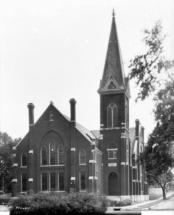The brick Gothic revival building has two gabled bays joined at a right angle. A square tower at the intersection is topped by a spire.