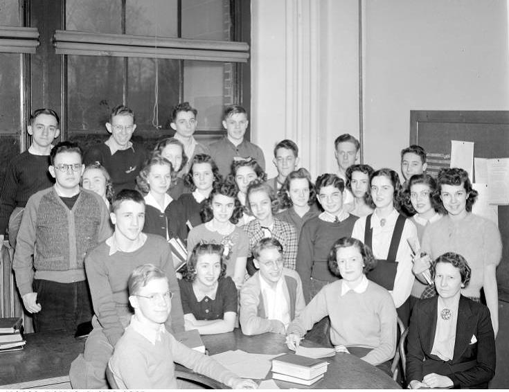 A group of about 30 people, mostly teenagers, gather around a desk in a classroom.
