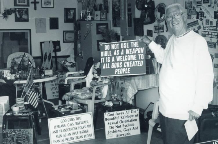 A man stands in a heavily decorated room. He is holding a sign that reads "Do not use the bible as a weaspon it is a welcome to all gods created people."