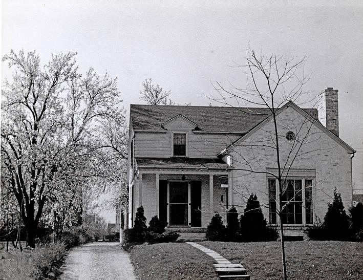 The house is a small, brick and clapboard house, with a covered porch left front and a bay window to the right. The peaked roof has brick chimney at one end.