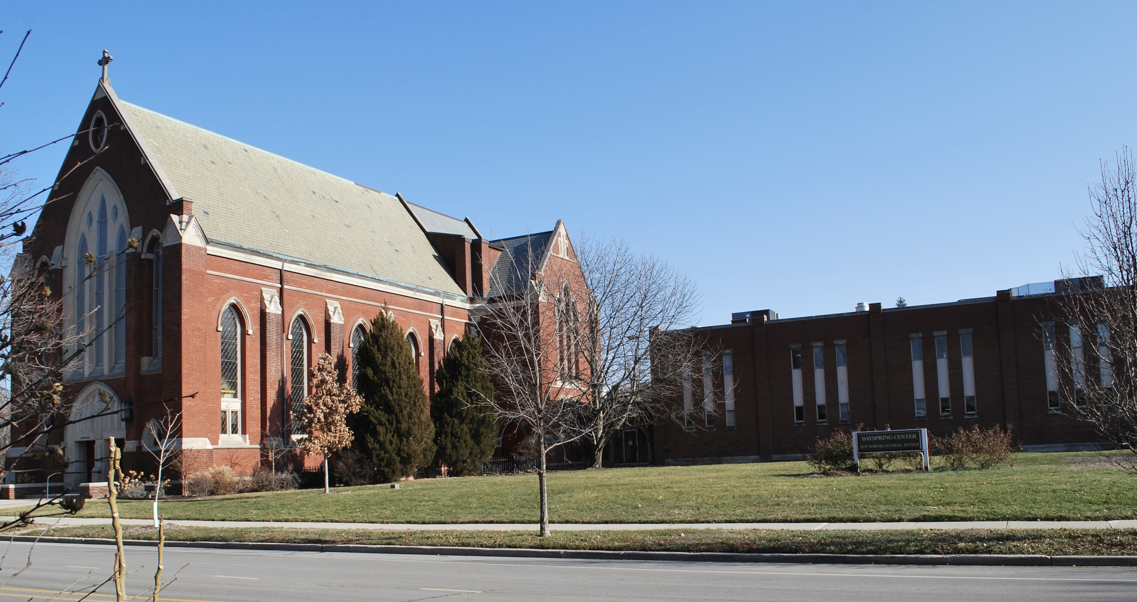 A rectangular brick building with several windows in sets of threes is attached to a large brick church with gabled roof. 