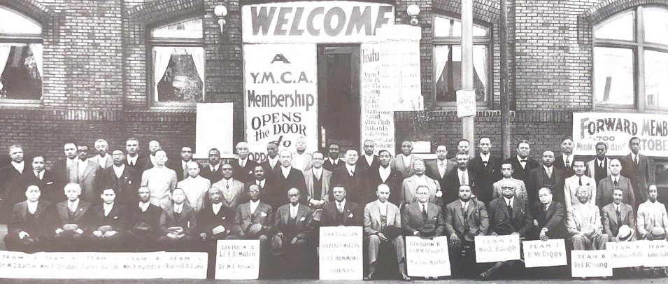 A group of men pose together in front of the YMCA building. Several men are holding signs.