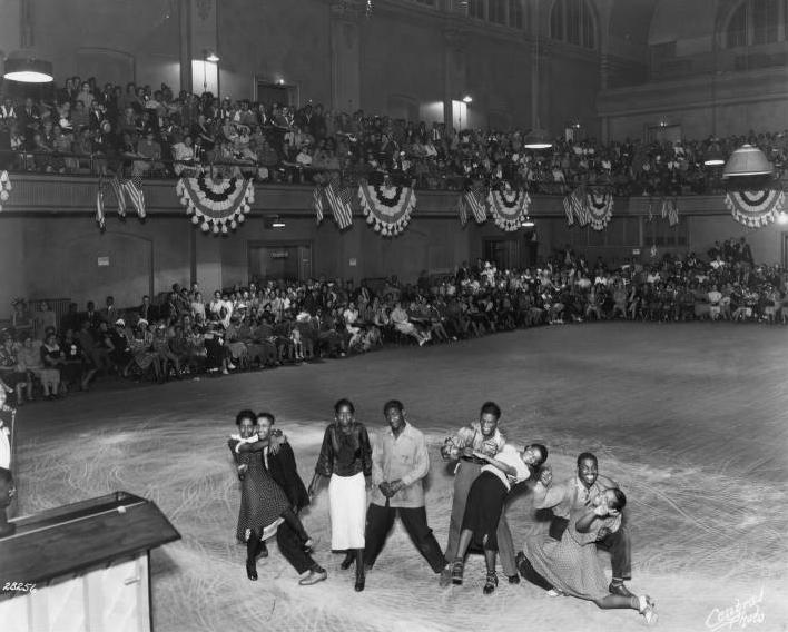 A group of teenagers dance while a crowd of people sit on the sides of the room and watch.