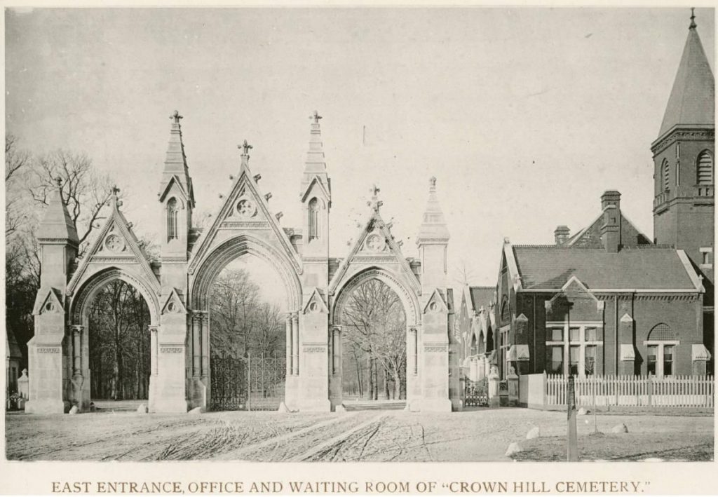 The gothic gateway is comprised of two slightly smaller gothic arches on either side of a center arch. Four small and pointed turret structures rise from the gate also. To the right is the brick, gothic revival residence and office.