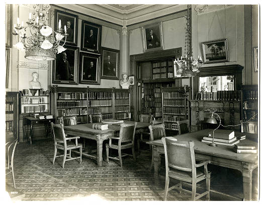The library is a high-ceiling room with wide crown molding. There are tables with chairs around them, several portraits on the walls, built-in bookcases and decorative chandeliers.