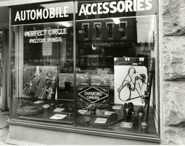 A glass storefront reads "Automobile Accessories. Perfect Circle Piston Rings" and displays several machine parts.