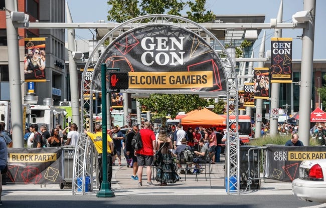 A metal arch with a banner reading "GenCon Welcom Gamers" is at the entrance to the block party held outside the convention center at GenCon. Many people are walking among the tables and colorful awnings and banners.