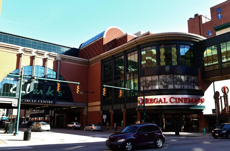 The movie theater entrance to the Circle Center Mall is rounded with windows filling multiple stories. A skybridge extends from the right side of the entrance and extending from the left side is a wing of the building elevated above the street. 