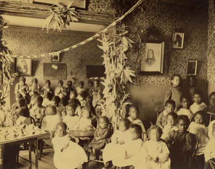 Several children are crowded in chairs and at tables in two adjoining rooms of a house.