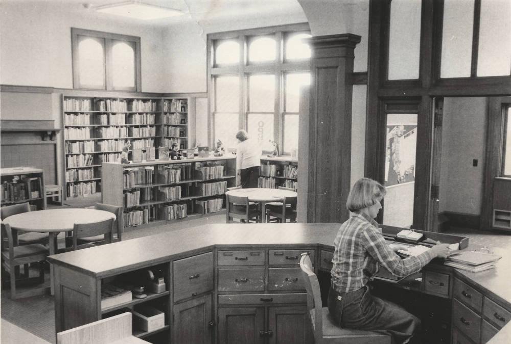 A woman sits behind a large wooden desk. Book-filled shelves line the walls in the background.