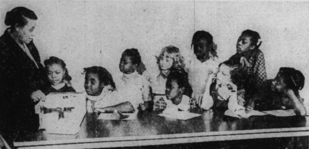 A group of children sit together and look towards a woman.