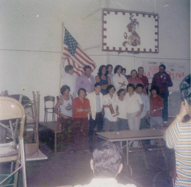 A group of young adults are gathered together for a group photograph. A banner featuring a latin american figure in traditional style. 