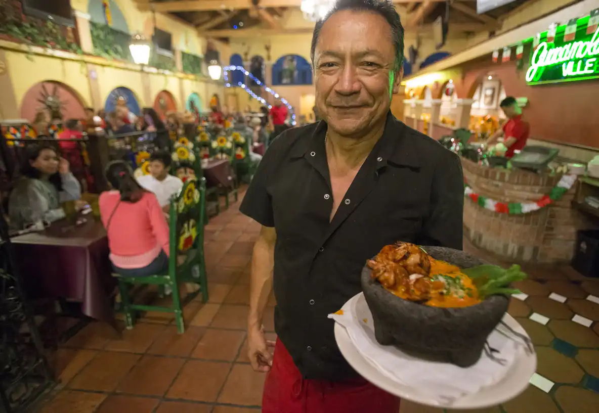 A man is holding a dish of food. In the background is a crowded, brightly colored Mexican restaurant.