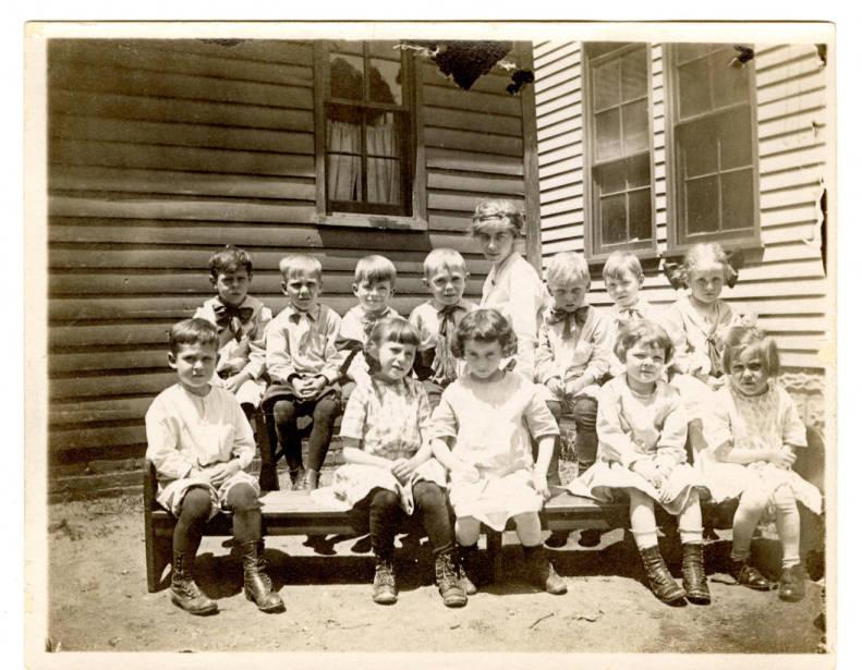 A group of children sit on two benches in front of a building.