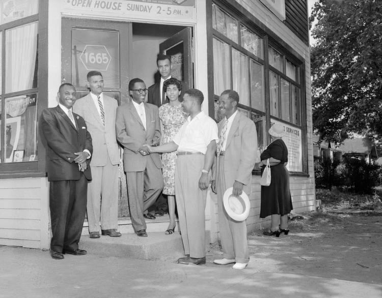 A small group of people stand at the entrance of a small building. Two men in the middle are shaking hands.