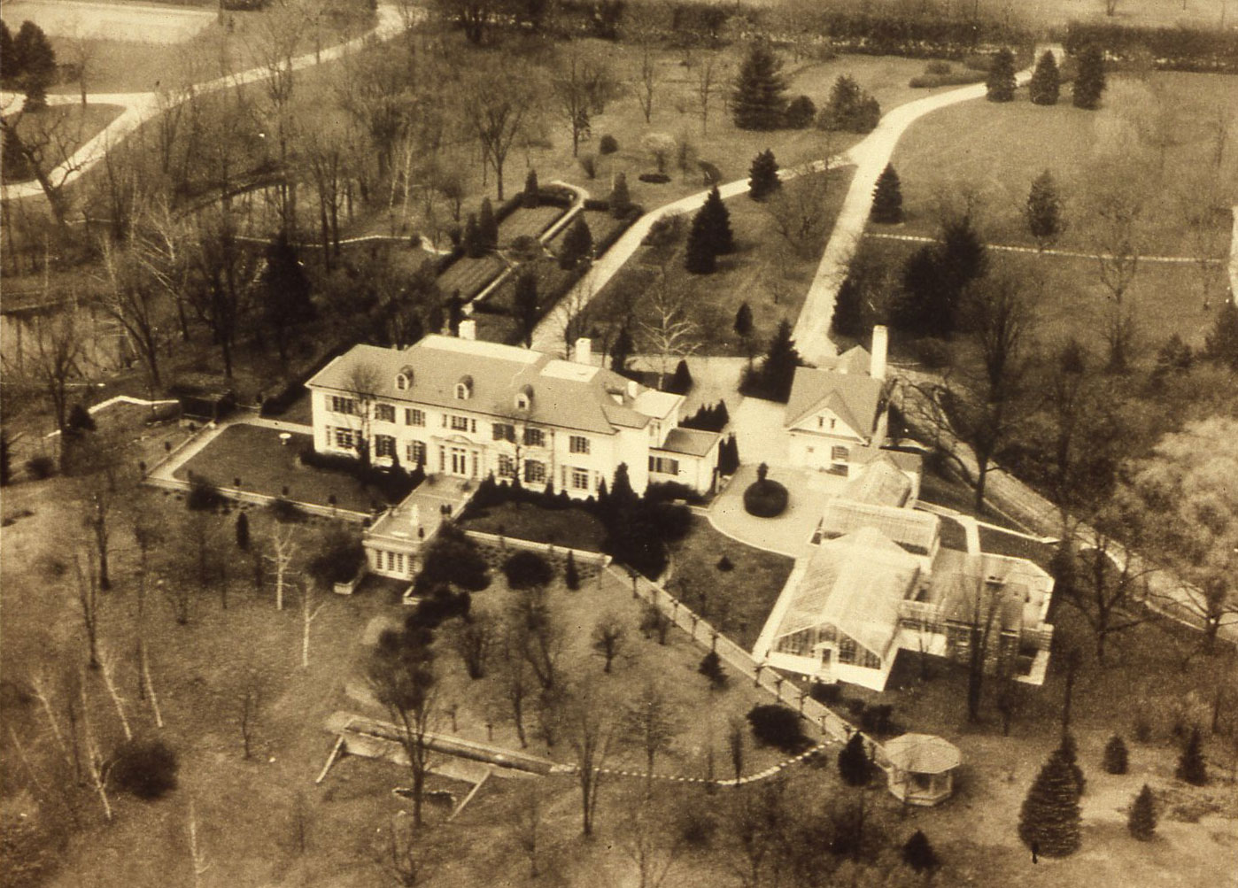Aerial view of a large, white, Colonial Revival mansion with a mansard roof set with dormer windows. There is a group of large greenhouses, a garage/servants building and a gazebo also on the estate.