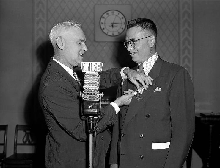 A man pins a medal on to Eugene C. Pulliam's jacket. Both men are standing behind a microphone. 