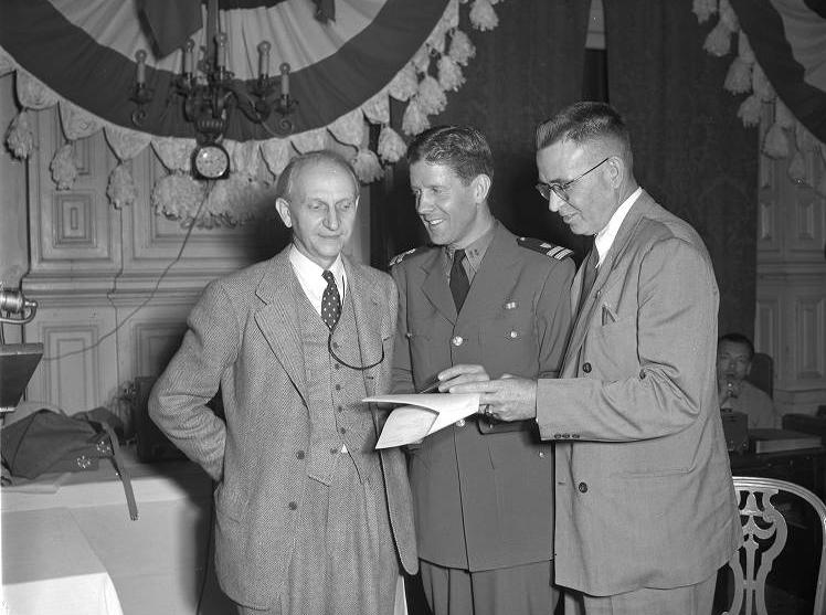 Three men stand together and look at a piece of paper. 