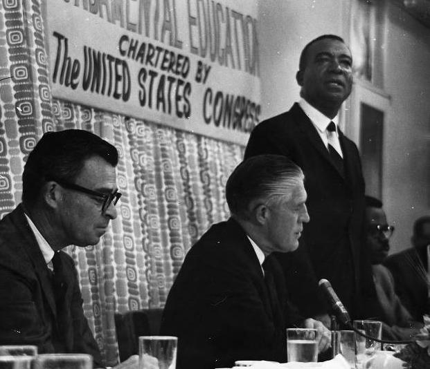 Two men sit and one man stands behind a table. A banner in the background reads "Fundamental Education chartered by the United States Congress."