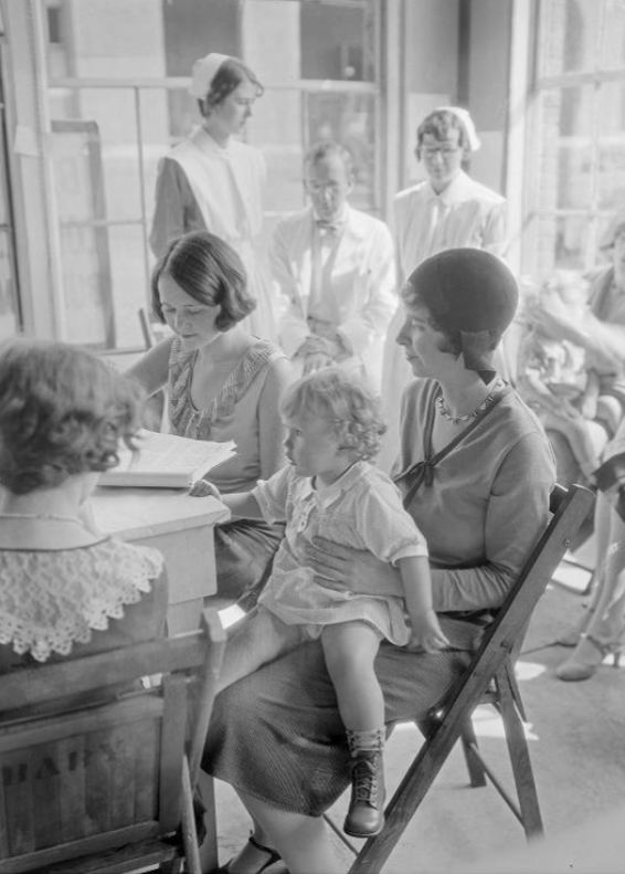 A woman sits at a table with a toddler on her lap. Two other women are at the table, and two nurses are in the background.