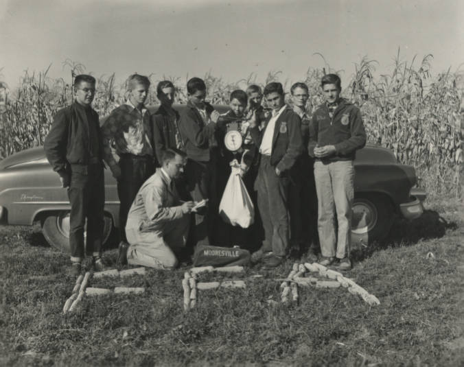 A group of young men stand around a man who is kneeling and weighing a bag on a scale. They are standing in front of a cornfield and have "FFA" spelled out in corncobs on the ground.
