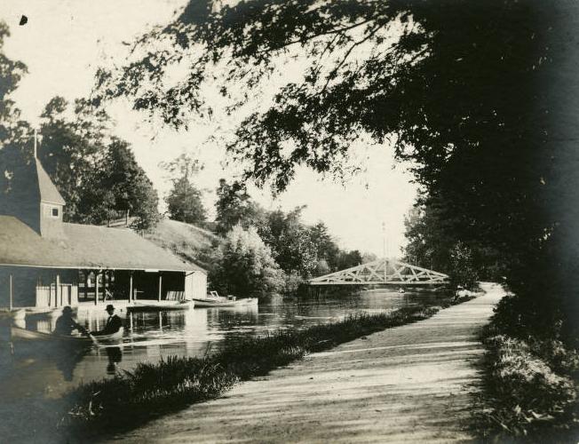 Two men paddle a canoe in the canal. The boathouse is on the left, the towpath on the right and a bridge crosses the canal ahead of them..