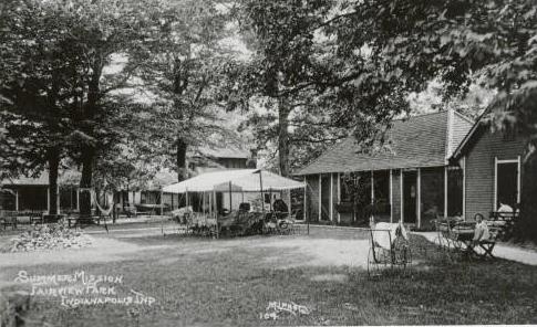 One-story cabins line two sides of a shaded yard. A large white canopy is erected on the grass.
