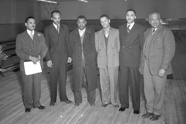 Six men stand in a semi-circle in a large room.