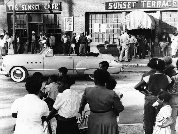 A large number of people stand on either side of a street. An empty convertible is parked in front of two entrances, one with the sign "The Sunset Cafe" over the door and the other with "Sunset Terrace" over its door.