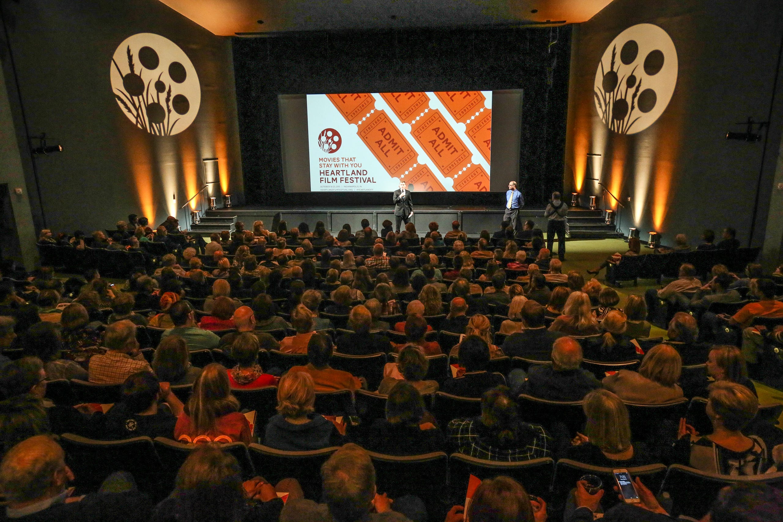 View from the back of a movie theater. The seats are filled with people. A person is talking at the front of the theater and the screen has graphics of tickets and the Heartland Film Festival logo. 