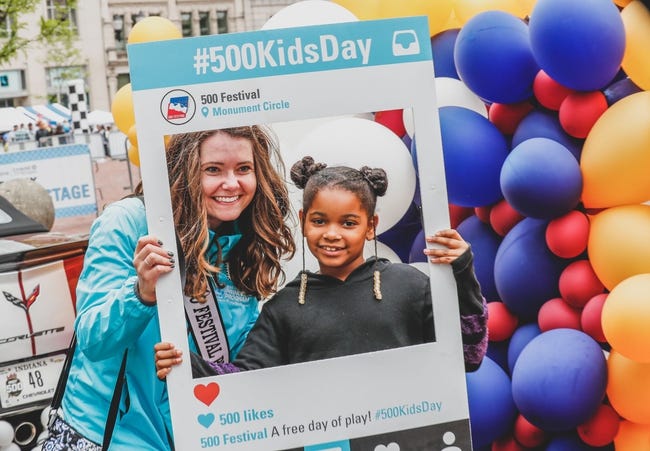 A young woman and a young girl hold a poster with a large square removed through which they can be photographed. The poster frame reads "#500KidsDay: 500 Festival Monument Circle." There are colorful balloons behind them.