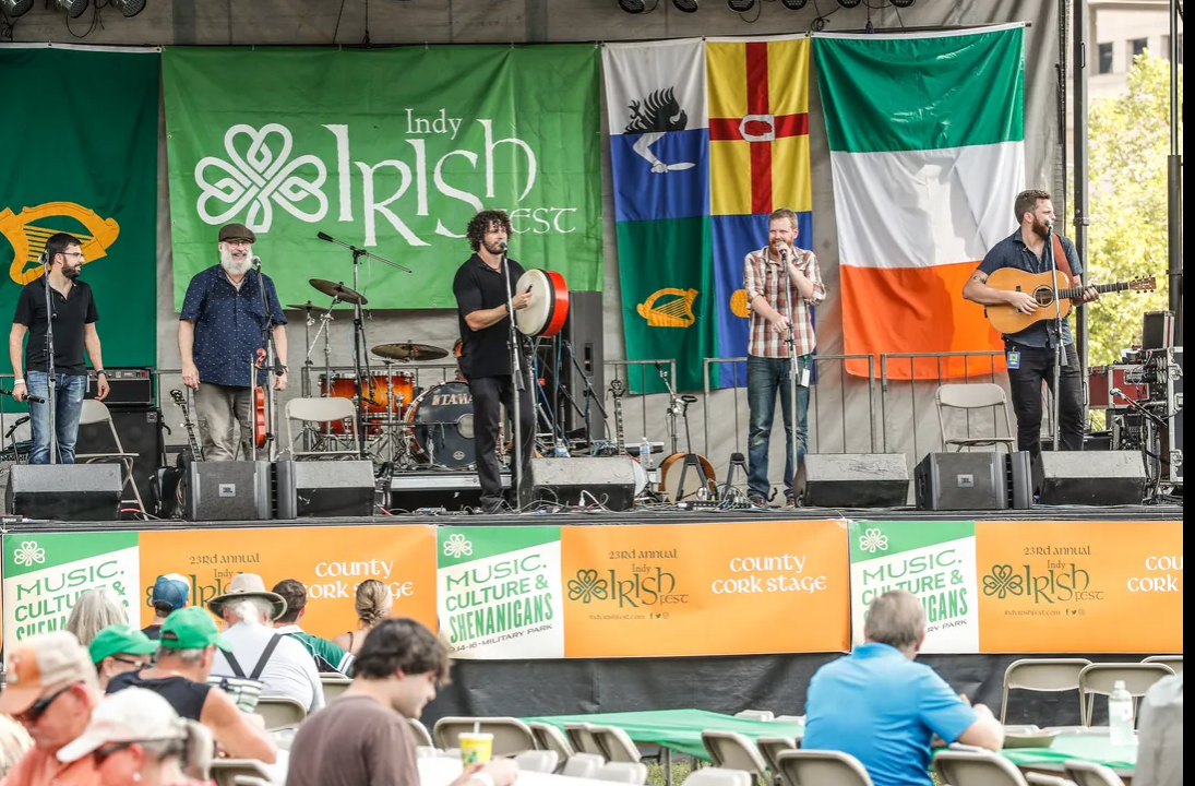 Five musicians perform on an outdoor stage. A large banner behind them has a shamrock and "Indy Irish Fest" on it. There are several other colorful flags and banners in the back and along the front edge of the stage. Long tables with people are in front of the stage.