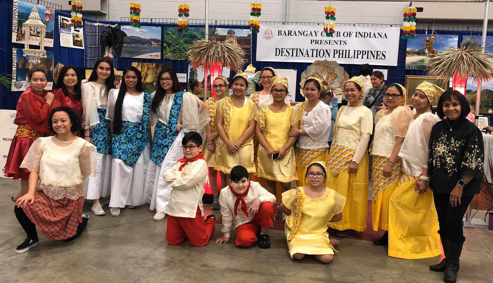 a group of people in various costumes pose in a room whose walls are filled with posters showing tropical destinations. Behind them, a banner reads "Barangay Club of Indiana Presents Destination Philippines".
