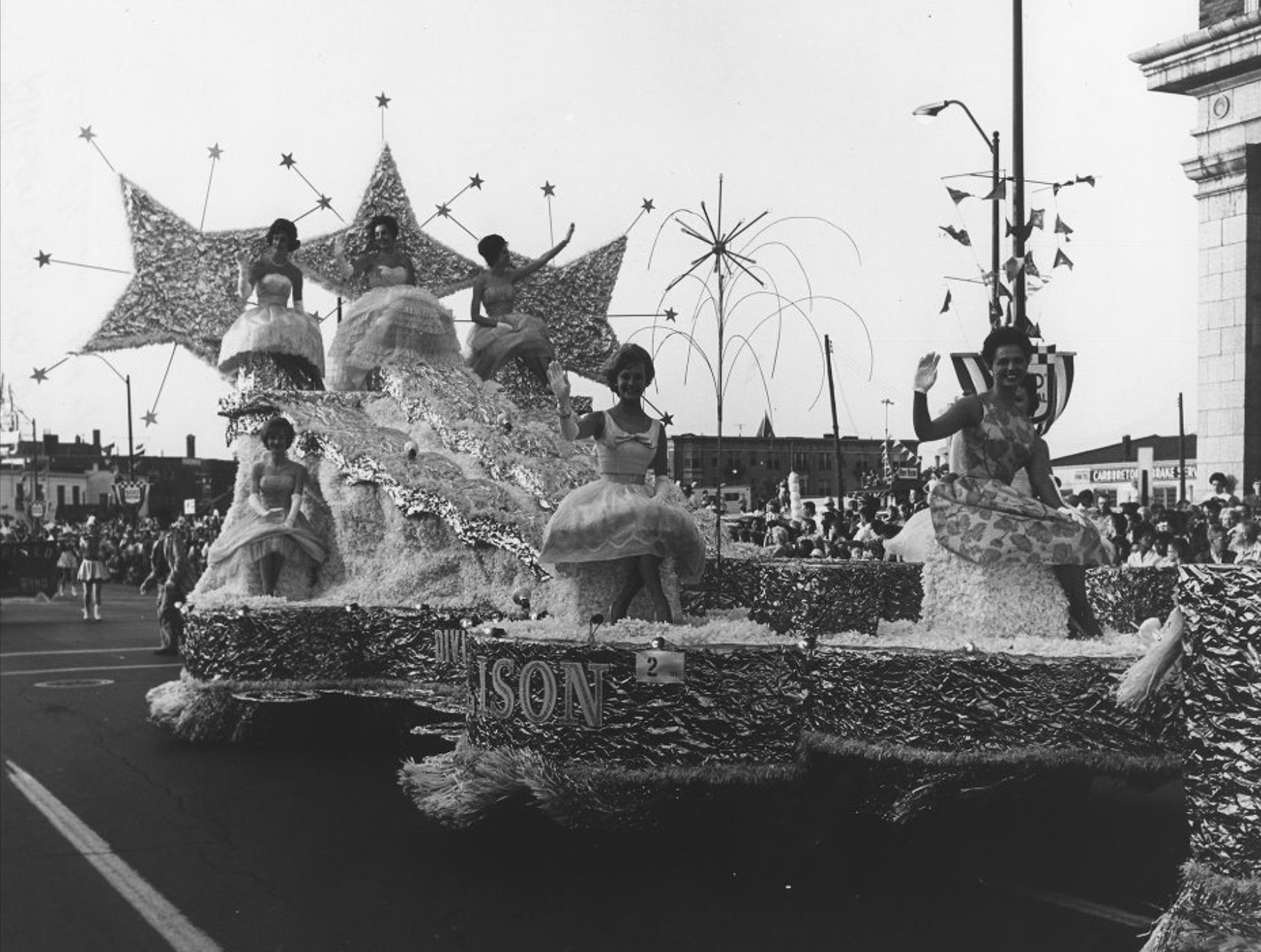 The parade float is decorated with large shooting stars. Several woman sit atop the float and wave at the crowds.