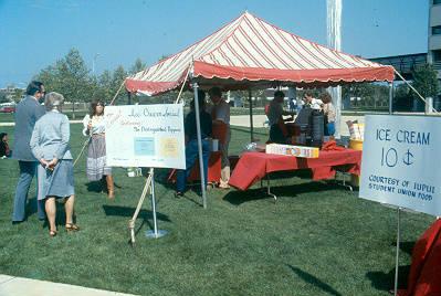 A couple tables are set up under a tent. Workers serve ice cream. A sign in the foreground reads "Ice Cream ten cents."