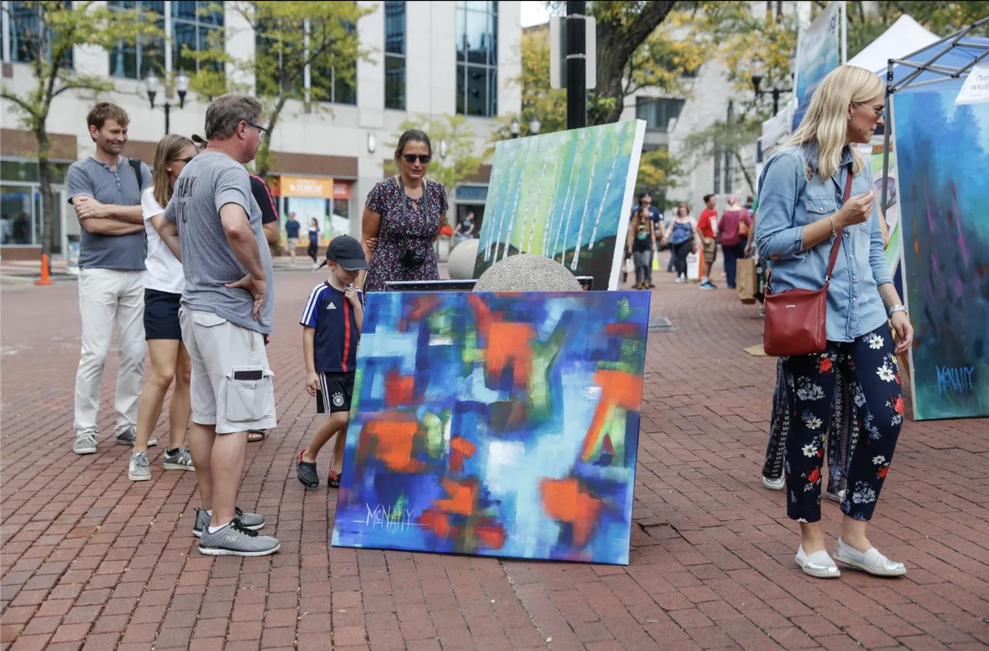 People look at several large paintings propped up on the brick sidewalk. The paintings are colorful and abstract.