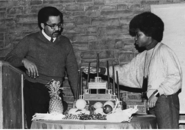 Two people stand on either side of a table. The table is covered with fruit and a candelabra.