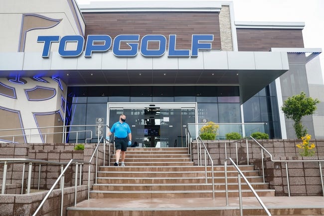 A man stands on the stone steps leading to the glassed entryway of a large, modern store. "TOPGOLF" is in large letters over the entrance.