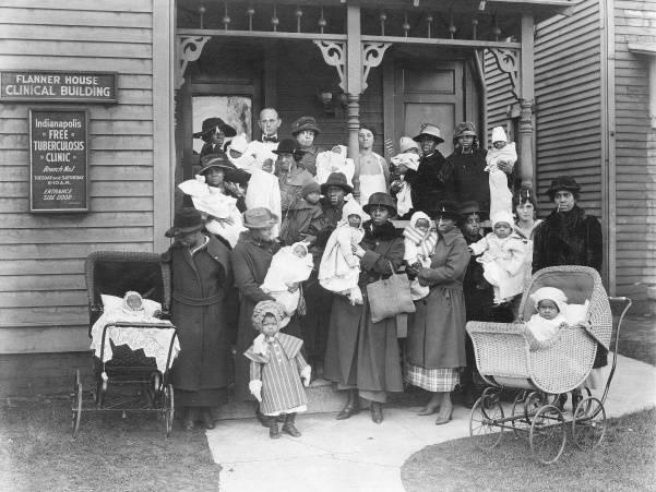 A group of women and their children stand together on the porch of a building with a sign on the front that reads "Flanner House Clinical Building."