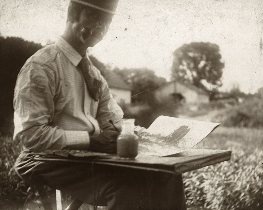 A younger Forsyth sits outside in a field. He is painting and holds a pipe in his mouth. 