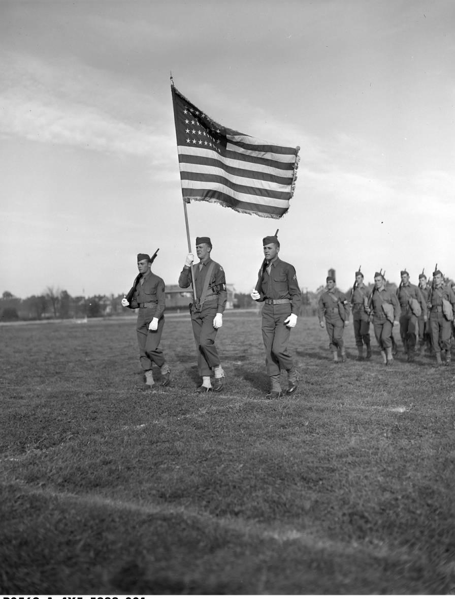 Several soldiers march in line. A soldier at the front holds an American flag.