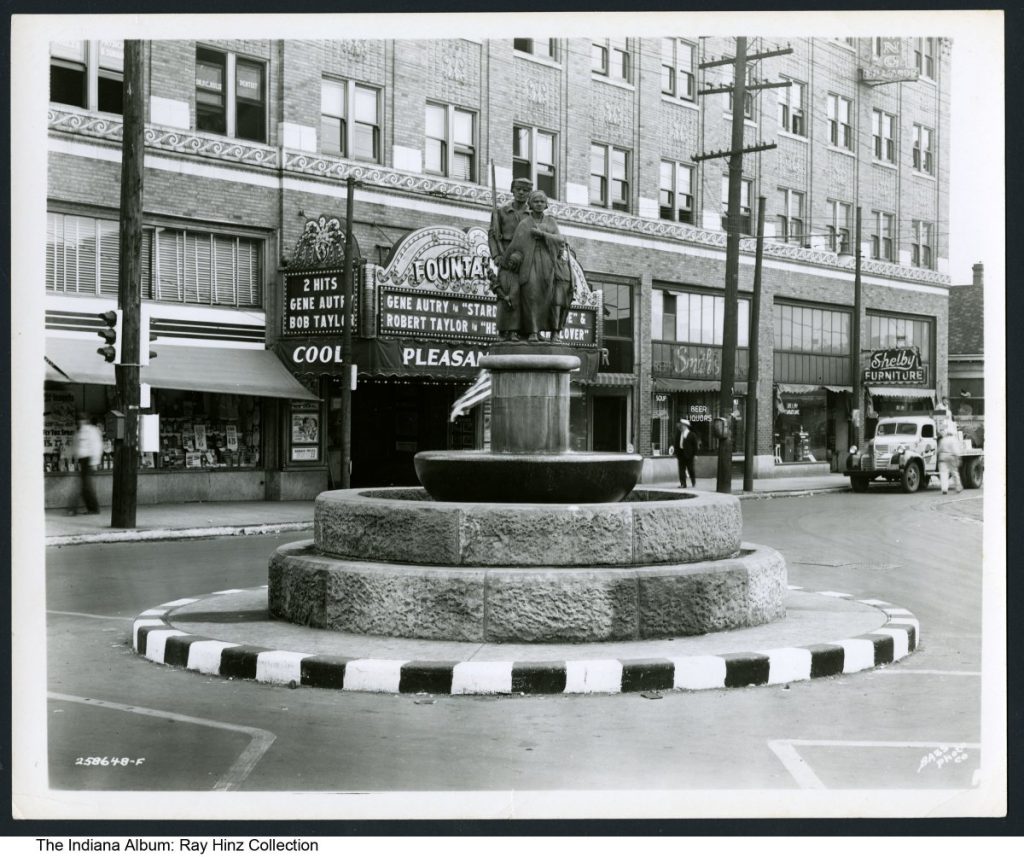 A statue depicting a man, woman, and child sits atop a fountain located in the middle of a street. In the background is the Fountain Square Theatre Building which includes Smith's Beer and Liquor store and Shelby Furniture.