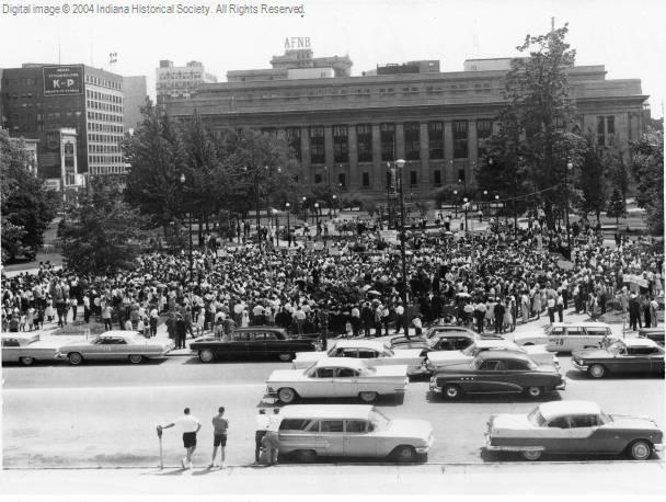 Crowds of people stand in front of a government building. Cars are driving down the street in the foreground.