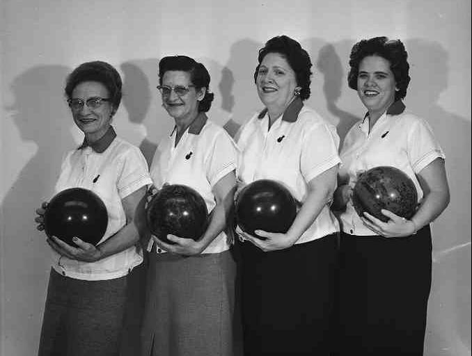 Four women wearing bowling shirts and holding bowling balls stand in a row.