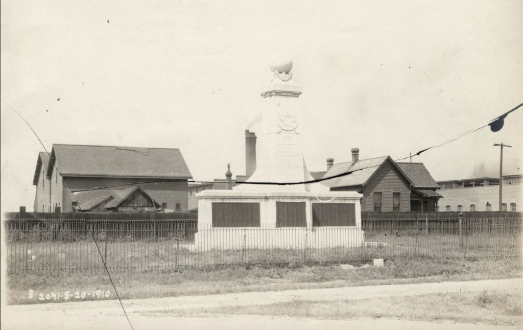 A monument sits in the middle of a small fenced in field. Buildings are in the background. 