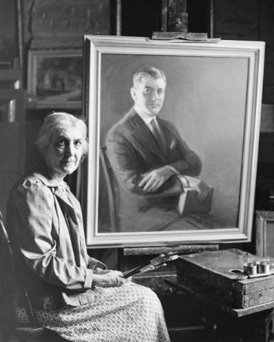 An older woman sits next to a painting of a man. She is holding several paint brushes. 