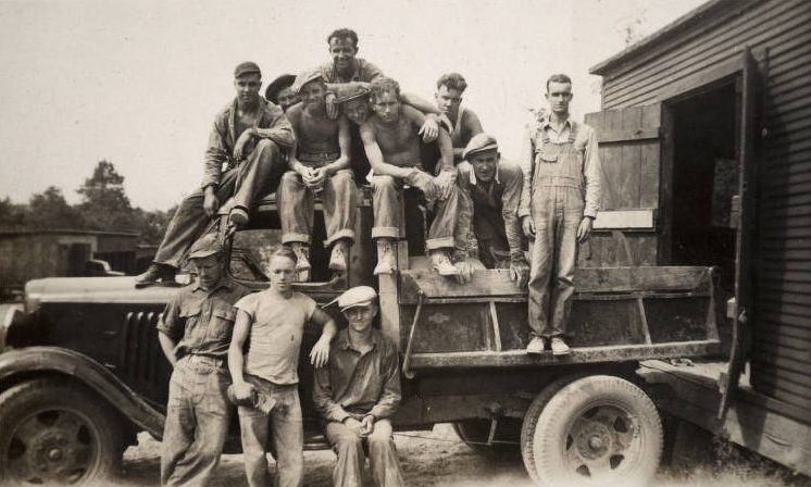 A group of men are on the roof, bed and running board of a military vehicle.
