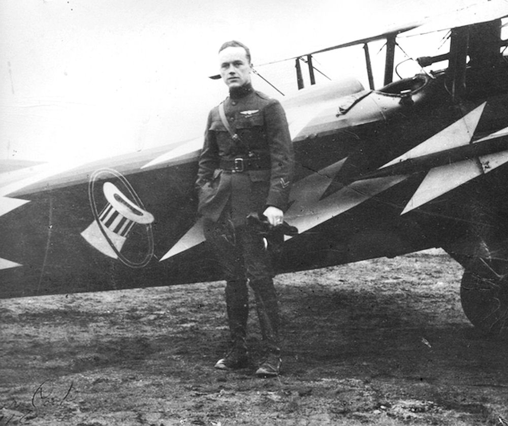 A man wearing a World War One flight lieutenant uniform stands next to an airplane.