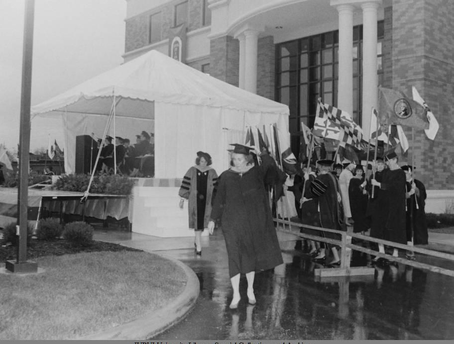 A tent is set up in front of a building. People sit in chairs under the tent. Outside of the tent, several people are holding up flags. Others are walking along a path. All people are wearing academic regalia.